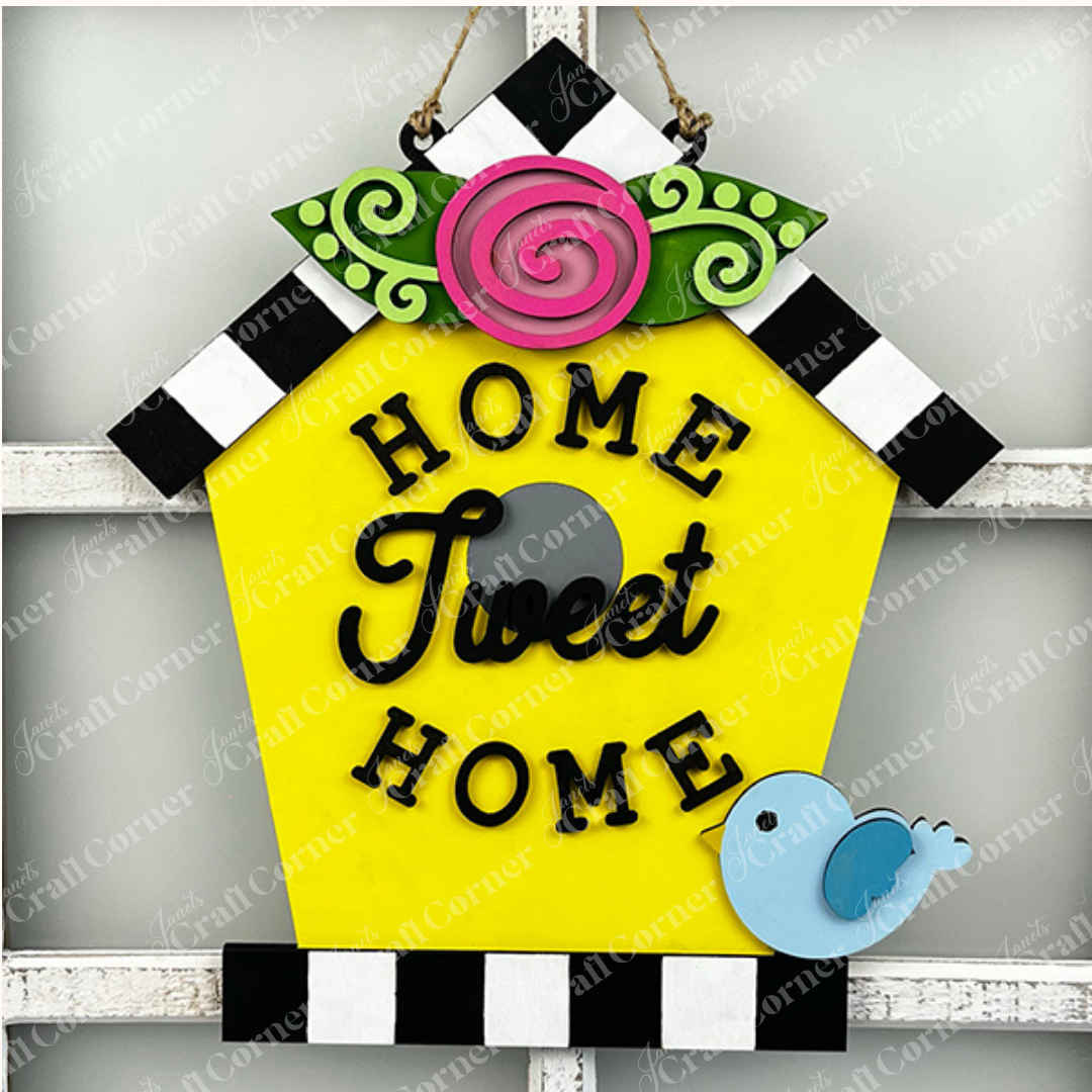 The Home Tweet Home Birdhouse Door Hanger by Janet's Craft Corner is a charming decorative piece featuring a yellow birdhouse sign with black and white striped roof edges. It is adorned with a pink rose and green leaves on top, making it the perfect addition to your home décor. The sign displays "Home Tweet Home" in black letters and includes a small blue bird in the bottom right corner.