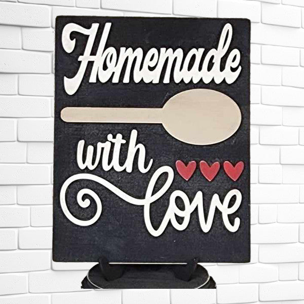 A sign from Janet's Craft Corner's Homemade Kitchen Interchangeable Sign DIY home decor craft kit is displayed against a white brick wall. It reads "Homemade with love" in white script, complemented by a charming wooden spoon graphic and three red hearts, adding a delightful touch to your DIY home decor.