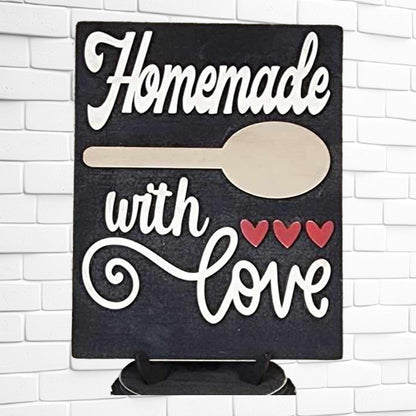 A sign from Janet's Craft Corner's Homemade Kitchen Interchangeable Sign DIY home decor craft kit is displayed against a white brick wall. It reads "Homemade with love" in white script, complemented by a charming wooden spoon graphic and three red hearts, adding a delightful touch to your DIY home decor.