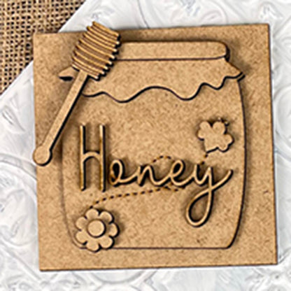 The Bee Swappable Squares from Janet's Craft Corner feature a honey jar design with "Honey" in cursive, plus flower and bee details, adding handmade charm to your decor on a white and burlap background.