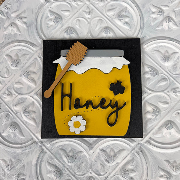 The Bee Swappable Squares from Janet's Craft Corner feature a yellow honey jar with a white lid, wooden dipper, white flower, and black bee on customizable squares. "Honey" is scripted in cursive on a black background for unique decor.