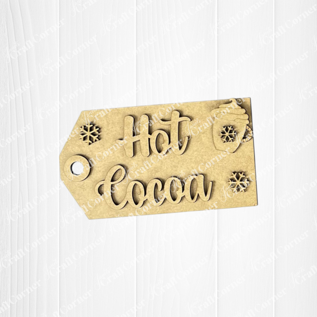 The Hot Cocoa Mini Shelf Sitter by Janet's Craft Corner features a tan tag with raised "Hot Cocoa" text, snowflakes, and a steaming cup—perfect as seasonal DIY home decor or craft kit. Set of 1, displayed on a white wood background.