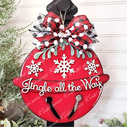 The Jingle Bell Door Hanger by Janet's Craft Corner adds a festive touch to your holiday decor with its red ornament adorned with white snowflakes and the phrase "Jingle all the Way" in cursive. It is completed with a black and white plaid bow, green foliage accents, and a backdrop of snow-dusted greenery.