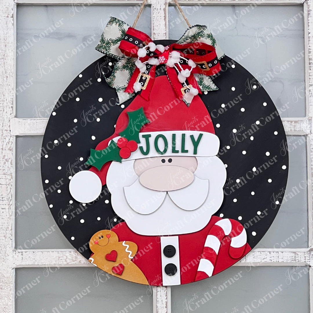 The Jolly Santa Round Sign from Janet's Craft Corner is a delightful holiday DIY home decor kit. It features Santa Claus wearing a "Jolly" hat set against a black polka dot backdrop, holding a gingerbread man and candy cane. A festive ribbon adorned with holly and bells completes this charming hand-painted decoration.