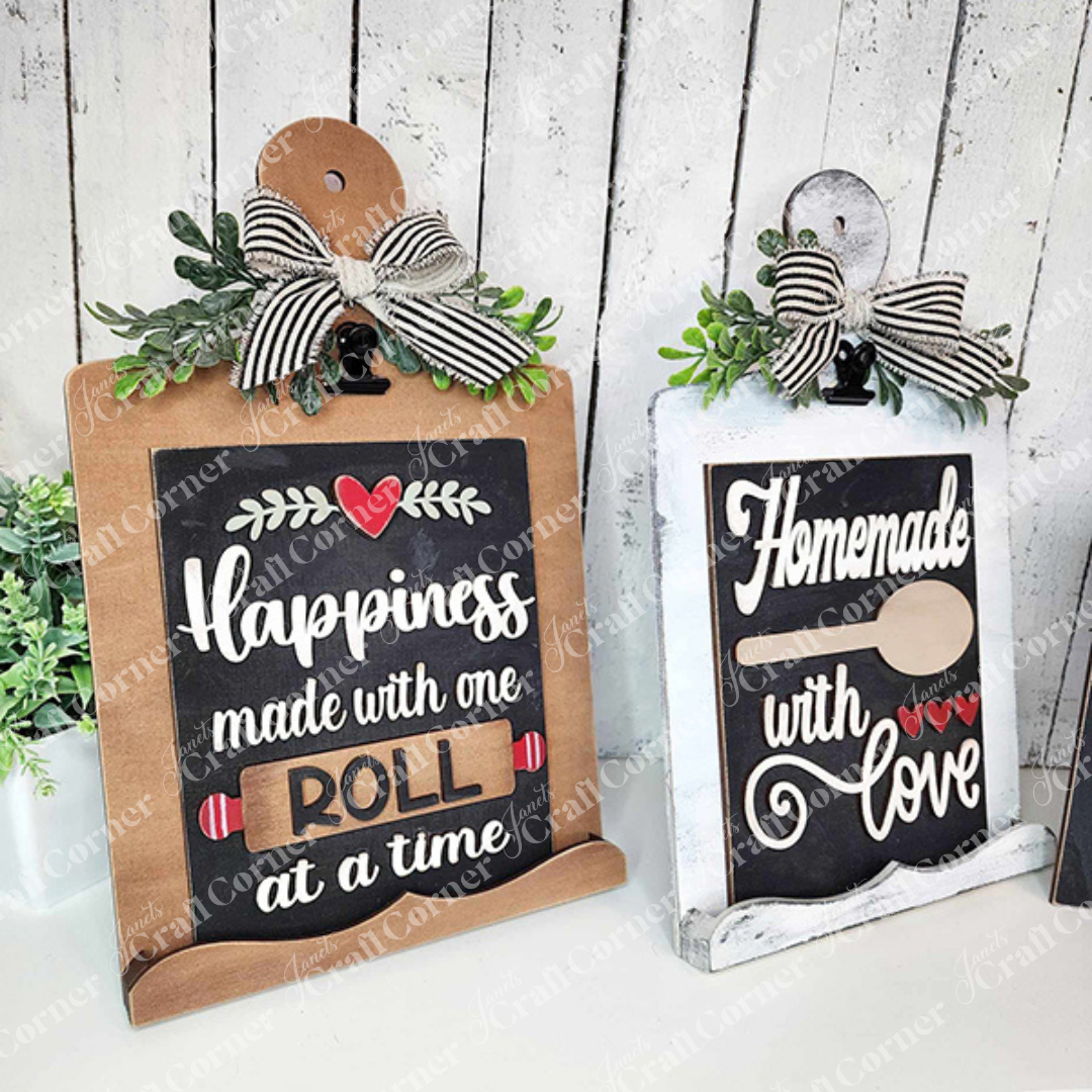 Janet's Craft Corner offers a Breadboard Sign Holder for Interchangeable Signs, perfect for farmhouse-inspired decor. This DIY home decor craft kit includes two charming designs: one reads "Happiness made with one roll at a time" featuring a rolling pin, and the other says "Homemade with Love" adorned with a spoon. Both signs are complemented by ribbon bows and greenery against a white wooden backdrop.