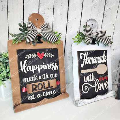 Janet's Craft Corner offers a Breadboard Sign Holder for Interchangeable Signs, perfect for farmhouse-inspired decor. This DIY home decor craft kit includes two charming designs: one reads "Happiness made with one roll at a time" featuring a rolling pin, and the other says "Homemade with Love" adorned with a spoon. Both signs are complemented by ribbon bows and greenery against a white wooden backdrop.