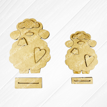 Janet's Craft Corner offers a Lamb Shelf Sitter DIY Kit featuring two charming wooden sheep cutouts with curly wool, facial details, a heart on each body, and bottom slots. One is larger than the other against a white background—ideal as home décor!.