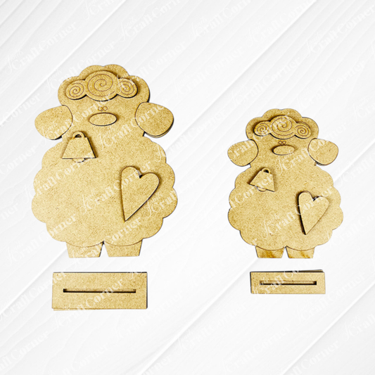 Janet's Craft Corner offers a Lamb Shelf Sitter DIY Kit featuring two charming wooden sheep cutouts with curly wool, facial details, a heart on each body, and bottom slots. One is larger than the other against a white background—ideal as home décor!.