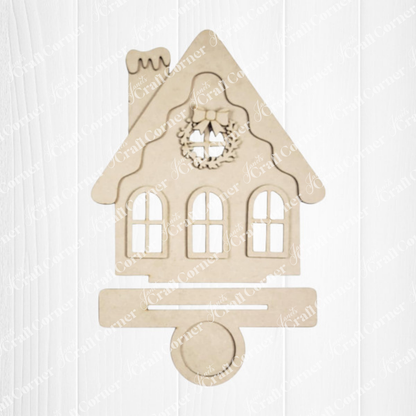 The Victorian Gingerbread House Votive Holder DIY craft kit by Janet's Craft Corner features a festive wooden cutout with a wreath above the windows and a slot at the bottom, ideal for seasonal decor, displayed on a white wood background.