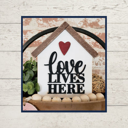 Introducing the "Love Lives Here Decor Set" by Janet's Craft Corner. This delightful decorative piece is designed in the shape of a house with a rustic wooden roof and an endearing red heart. It features the phrase "Love Lives Here" elegantly displayed in black text. Ideal for enhancing your home décor, it fits beautifully among greenery and wooden beads on your tiered tray, providing a charming contrast against a brick wall backdrop.