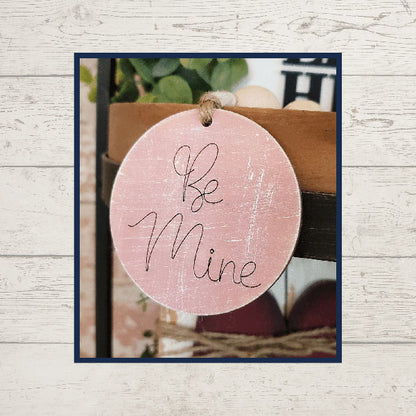 A round, pink wooden tag displaying "Be Mine" in an elegant cursive font is a charming addition to any tiered tray setup. Hanging by a string against a blurred backdrop, it enhances rustic surfaces and greenery accents, making it the ideal piece for home décor enthusiasts. Discover this beautiful item as part of Janet's Craft Corner's Love Lives Here Decor Set DIY home decor craft kit.