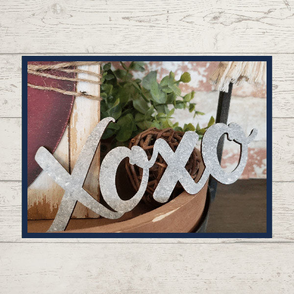 The Love Lives Here Decor Set from Janet's Craft Corner features a charming tiered tray display, which includes a metal sign inscribed with "Xoxo" on a wooden surface. This delightful arrangement is complemented by a green plant, a woven ball, and a wooden board, all set against a light, textured backdrop—making it an appealing addition to any home décor.