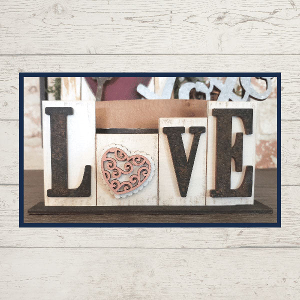 The Love Lives Here Decor Set from Janet's Craft Corner features a charming wooden block arrangement that spells out "LOVE." The letter "O" is cleverly replaced by a pink heart with an intricate swirling pattern, making it perfect for enhancing your tiered tray décor. It all comes together against a rustic wood-textured wall, creating a cozy home atmosphere.