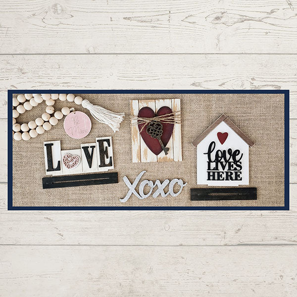 The "Love Lives Here Decor Set" by Janet's Craft Corner is a delightful DIY home decor craft kit. It includes a rustic collage perfect for a tiered tray, featuring the word "LOVE" with wooden beads, a small plaque showcasing a heart and key design, the phrase "Xoxo," and a charming wooden house shape with the text "Love lives here." All elements are beautifully arranged on burlap against a white wood background.