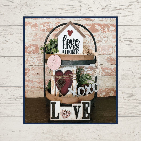 A charming home décor display features the "Love Lives Here Decor Set" from Janet's Craft Corner, showcasing a "Love Lives Here" sign, potted plants, a red heart, and the words "Xoxo" and "Love" in various fonts. This delightful arrangement adorns a tiered tray against a textured brick wall.
