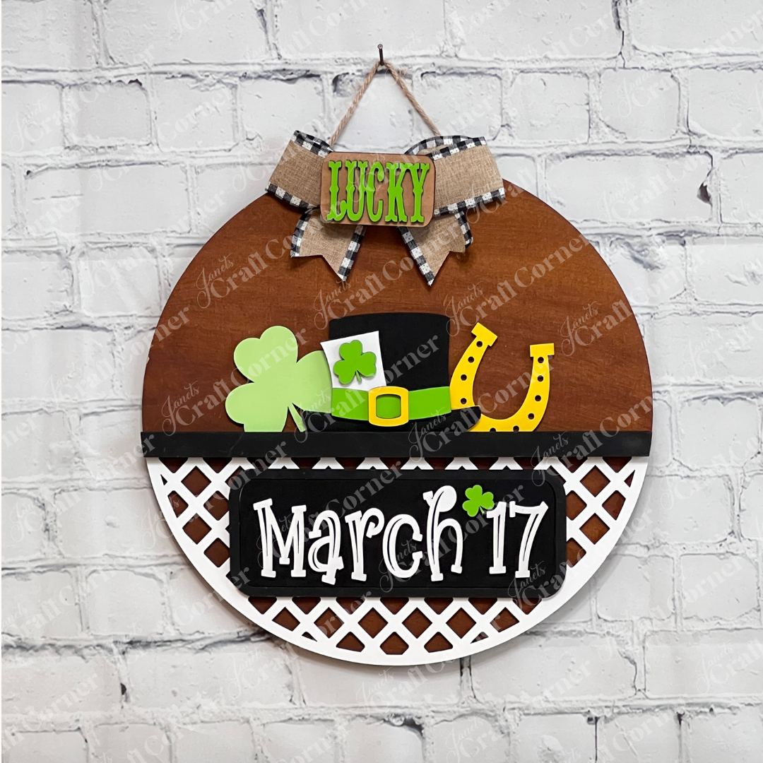 A decorative white fence display against a brick wall features interchangeable inserts from Janet's Craft Corner celebrating St. Patrick's Day, including a green shamrock, a black hat with a shamrock, and a yellow horseshoe. The plaque reads "March 17" with a small shamrock accent.