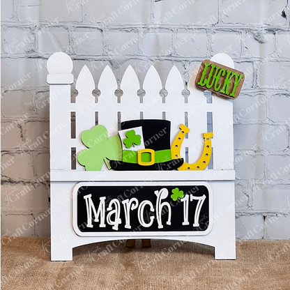 A decorative white fence display against a brick wall features interchangeable inserts from Janet's Craft Corner celebrating St. Patrick's Day, including a green shamrock, a black hat with a shamrock, and a yellow horseshoe. The plaque reads "March 17" with a small shamrock accent.