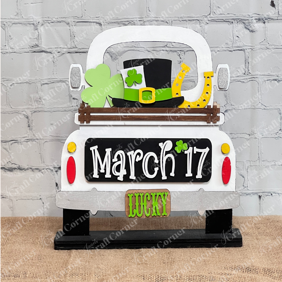 The "March 17 Interchangeable Insert" from Janet's Craft Corner is a decorative piece designed to resemble the back of a truck, perfect for celebrating St. Patrick's Day. It features symbols like a shamrock, leprechaun hat, and horseshoe, along with the date "March 17." With "LUCKY" displayed on the license plate, this DIY home decor kit makes an ideal piece of interchangeable home decor against a white brick wall.