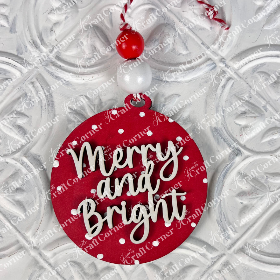 This charming piece of home décor from Janet's Craft Corner, included in the Round Christmas Ornaments | DIY Decoration Kits, features festive "Merry and Bright" text in white cursive on a red background with polka dots. It comes attached to a string adorned with red and white beads and placed against a textured white surface, making it perfect for your crafting projects!