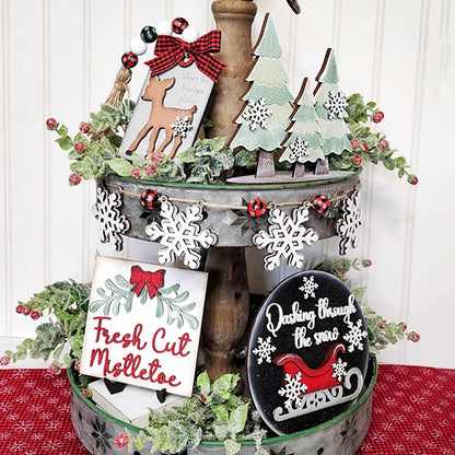 A festive Mistletoe Tiered Tray from Janet's Craft Corner, adorned with holiday decorations including evergreen trees, snowflakes, and a reindeer ornament. The set includes charming signs reading "Fresh Cut Mistletoe," as well as surrounding greenery and berries to complete the Christmas theme.