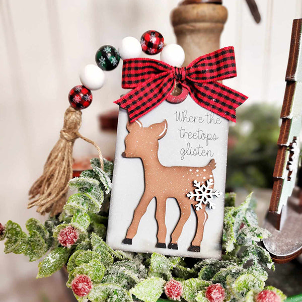 This festive DIY decor kit from Janet's Craft Corner, perfect for a mistletoe tiered tray, includes a wooden tag with a reindeer silhouette and snowflake cutout. It is embellished with a red and black plaid bow and beaded tassel. The text on the tag reads "Where the treetops glisten," surrounded by frosted greenery and tiny red berries.