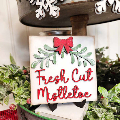 A charming piece from Janet's Craft Corner, the Mistletoe Tiered Tray DIY Decor Kit includes a decorative sign with a red bow and green mistletoe accents that spells "Fresh Cut Mistletoe" in vibrant red letters. Ideal for enhancing your holiday decor, this kit allows the sign to rest beautifully on a tiered tray, complete with festive Christmas-themed decorations.