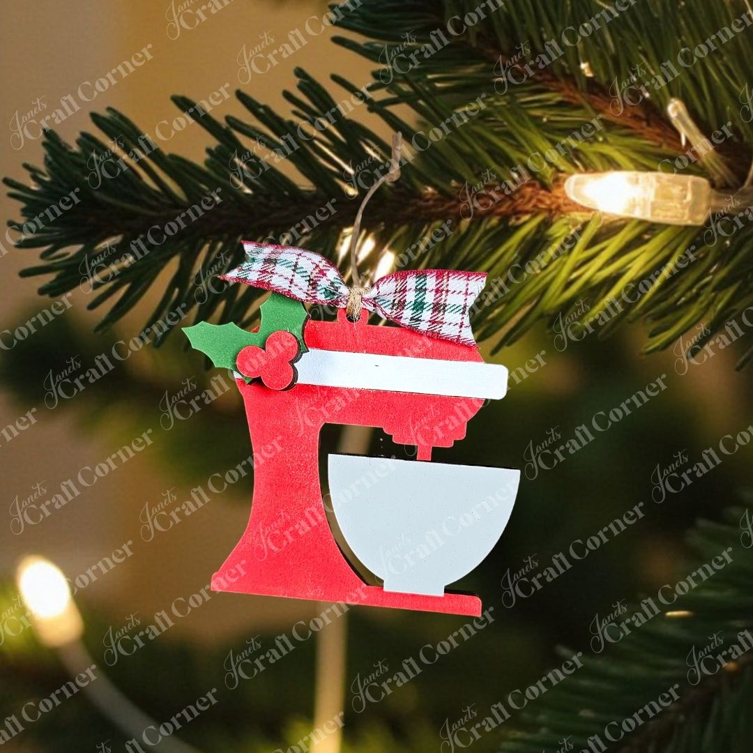 A hand-painted ornament, featuring a classic stand mixer with a bowl and adorned with holly and a plaid ribbon, hangs gracefully against a green pine background complete with pinecones. This charming piece from Janet's Craft Corner, known as the "Baking Spirits Bright Ornaments - Individual Painted," beautifully enhances your festive décor.