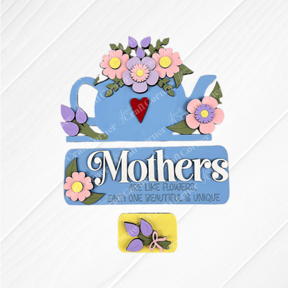 A decorative sign from Janet's Craft Corner, named "Mothers Interchangeable Insert - Painted," features a textured fabric with the phrase "Mothers are like flowers, each one beautiful & unique" in white lettering on a blue background. It is enhanced by a hand-painted teapot decor with pink floral details above and complemented by a small yellow rectangle with purple leaves below.