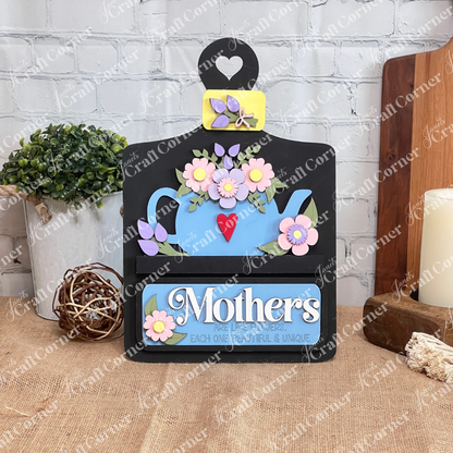 A delightful display piece from Janet's Craft Corner, the "Mothers Interchangeable Insert - Painted" on a decorative sign shaped like a breadboard. It features a charming teapot motif adorned with colorful flowers on its roof, against which the phrase "Mothers are like flowers, each one beautiful & unique" is elegantly displayed. The sign sits gracefully on a wooden surface with a brick wall providing the perfect backdrop.