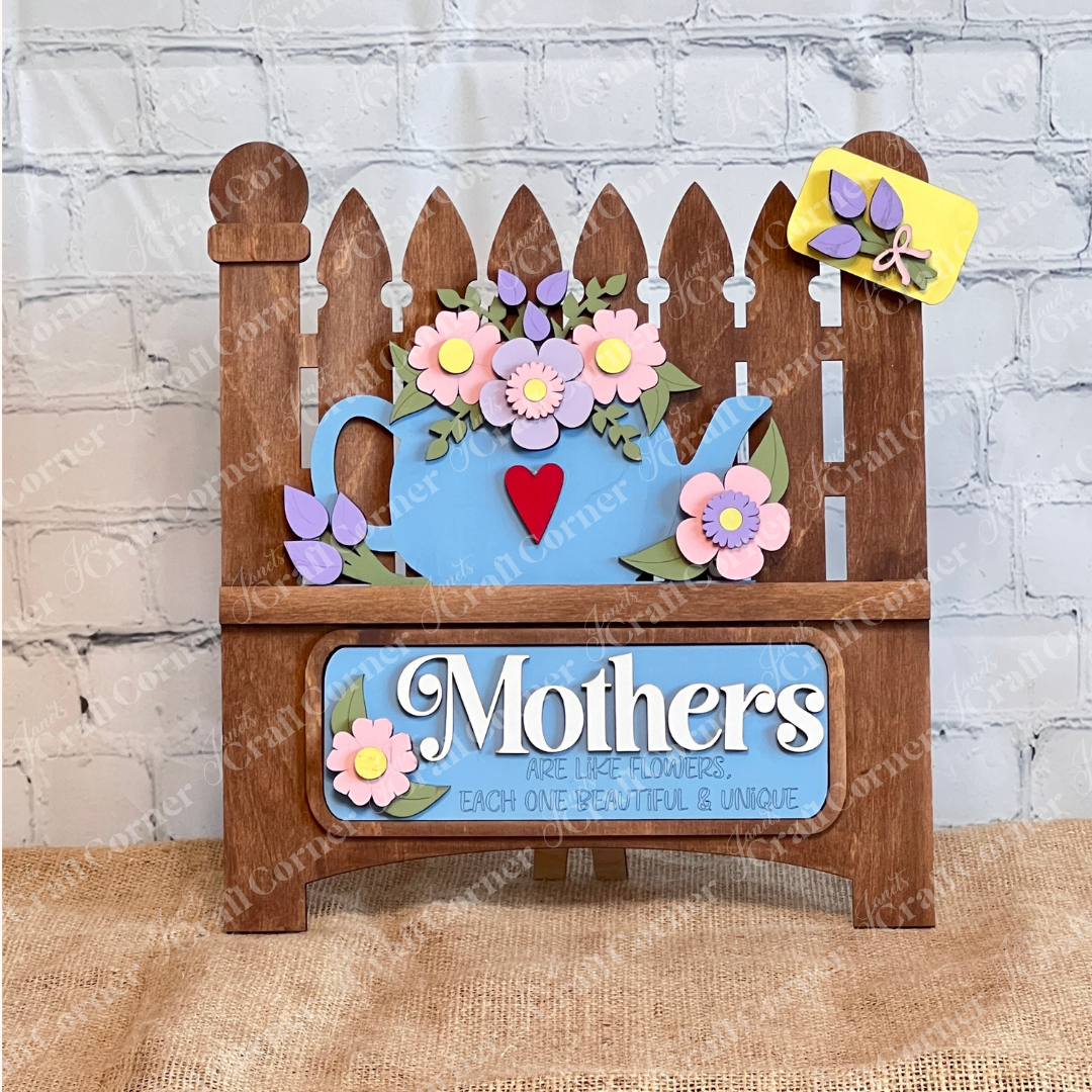 A delightful display piece from Janet's Craft Corner, the "Mothers Interchangeable Insert - Painted" on a decorative sign shaped like a garden fence. It features a charming teapot motif adorned with colorful flowers on its roof, against which the phrase "Mothers are like flowers, each one beautiful & unique" is elegantly displayed. The sign sits gracefully on a wooden surface with a brick wall providing the perfect backdrop.