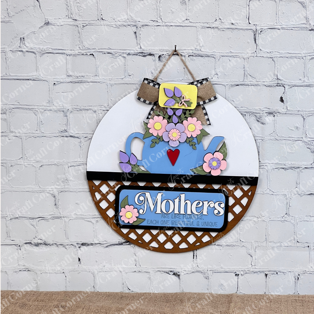 A delightful display piece from Janet's Craft Corner, the "Mothers Interchangeable Insert - Painted" on a decorative sign shaped like a door hanger. It features a charming teapot motif adorned with colorful flowers on its roof, against which the phrase "Mothers are like flowers, each one beautiful & unique" is elegantly displayed. The sign sits gracefully on a wooden surface with a brick wall providing the perfect backdrop.