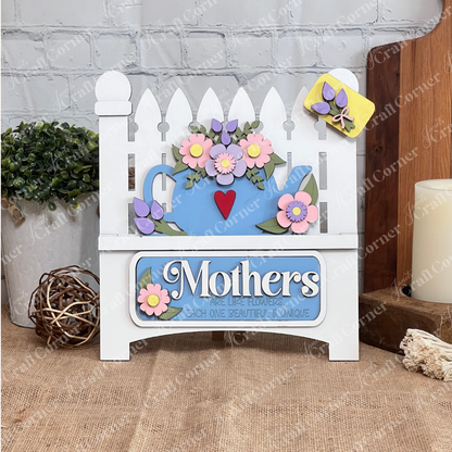 A delightful display piece from Janet's Craft Corner, the "Mothers Interchangeable Insert - Painted" on a decorative sign shaped like a picket fence. It features a charming teapot motif adorned with colorful flowers on its roof, against which the phrase "Mothers are like flowers, each one beautiful & unique" is elegantly displayed. The sign sits gracefully on a wooden surface with a brick wall providing the perfect backdrop.