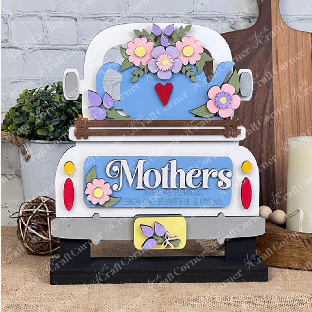 A delightful display piece from Janet's Craft Corner, the "Mothers Interchangeable Insert - Painted" on a decorative sign shaped like a vintage truck. It features a charming teapot motif adorned with colorful flowers on its roof, against which the phrase "Mothers are like flowers, each one beautiful & unique" is elegantly displayed. The sign sits gracefully on a wooden surface with a brick wall providing the perfect backdrop.