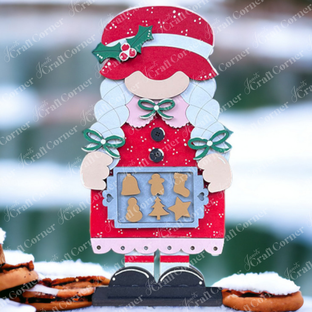 Decorate your home with Janet's Craft Corner's Mrs Claus Baking Gnome Shelf Sitter. This DIY seasonal décor kit includes a Mrs Claus in a red dress holding holiday cookies, set against a snowy background with blurred greenery and cookies stacked in the foreground.