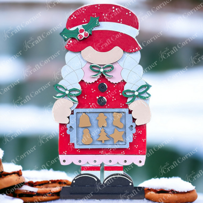Decorate your home with Janet's Craft Corner's Mrs Claus Baking Gnome Shelf Sitter. This DIY seasonal décor kit includes a Mrs Claus in a red dress holding holiday cookies, set against a snowy background with blurred greenery and cookies stacked in the foreground.