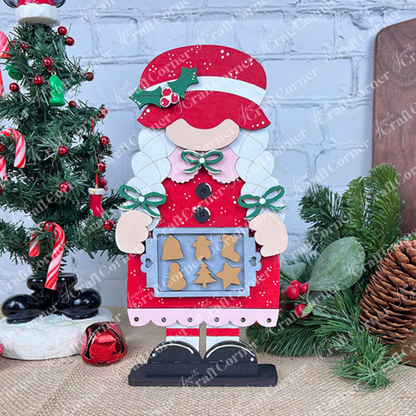 A festive Janet's Craft Corner Mrs Claus Baking Gnome, adorned in red, stands holding a tray filled with cookies shaped like trees, stars, and gingerbread people. It is surrounded by Christmas decorations featuring a DIY kit tree embellished with baubles and candy canes, all against a brick wall backdrop.