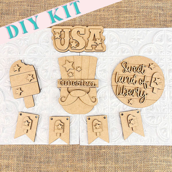 Flat lay of Janet's Craft Corner's Patriotic - Tiered Tray Kit, featuring wooden cutouts like "USA," a popsicle, and "Sweet Land of Liberty." Ideal for crafting patriotic tiered tray displays, it comes with star-themed banner pieces. Set against decorative burlap and tile, this farmhouse-style decor piece celebrates American spirit.