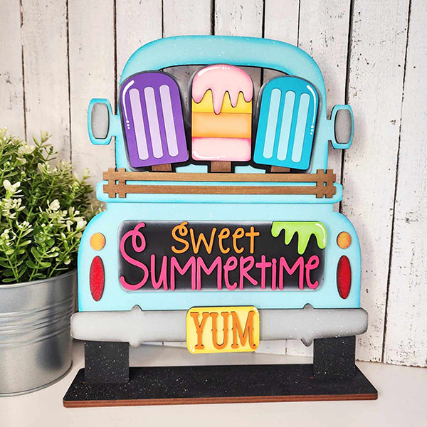 Add a charming touch to your home décor with Janet's Craft Corner's DIY home decor kit, featuring the Popsicle Interchangeable Insert. This decorative piece showcases a blue truck with vibrant popsicles and "Sweet Summertime" elegantly written in pink and orange above a yellow "YUM" license plate. It’s perfect for placing next to a potted plant on a wooden surface.