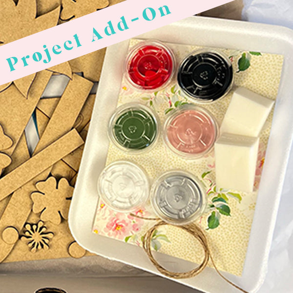 Wood cut-out kit from Janet's Craft Corner, named Project Kit Add-On, includes pre-cut wooden pieces, a spool of twine, and small containers of red, black, green, pink, white, and silver paints set on floral-patterned paper. The top is adorned with the Project Add-On label.