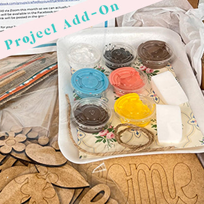 Art and craft supplies from Janet's Craft Corner are neatly arranged on a table, featuring small containers of paint, sponges, twine, pre-cut wooden pieces, and a decorative tray. A label reads "Project Kit Add-On.