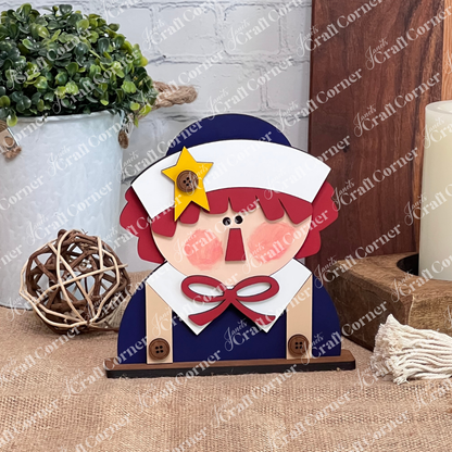 The Raggedy Ann & Andy Shelf Sitters DIY craft kit by Janet's Craft Corner features a red-haired wooden figure with a white bonnet, yellow star, buttons, and bow—sitting on burlap with rustic decor, perfect for fans and seasonal home decor.