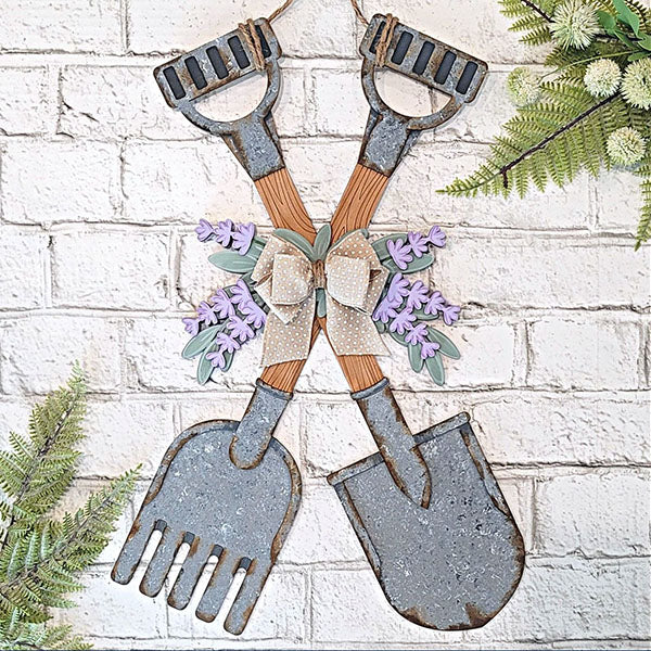 Introduce a touch of rustic elegance to your space with Janet's Craft Corner's Shovel and Rake Door Hanger. This delightful decor piece features two crossing garden tools, a shovel and rake, both with wooden handles. Decorated with lavender flowers and an eye-catching polka dot ribbon, it perfectly complements a white brick wall backdrop amidst vibrant green foliage.