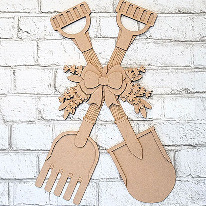 The Shovel and Rake Door Hanger by Janet's Craft Corner features two crossed gardening tools against a white brick wall, with bows and leaves accents. This DIY craft kit is ideal for a paint-it-yourself project or as a charming addition to rustic wood decor.