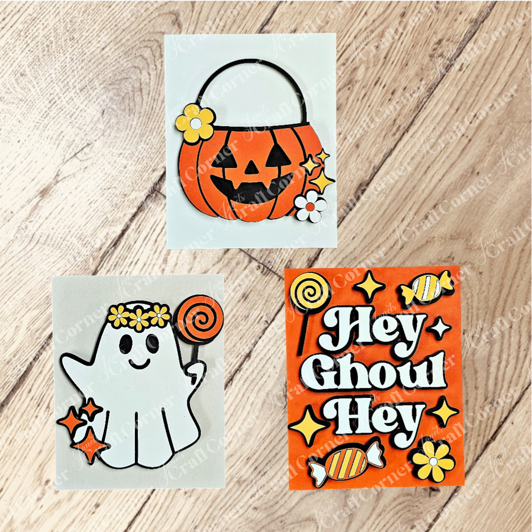 Janet's Craft Corner’s Halloween Farmhouse Signs set features three festive pieces: a jack-o’-lantern pail with flowers, a ghost with flower crown and lollipop, and an orange “Hey Ghoul Hey” sign with candy accents.