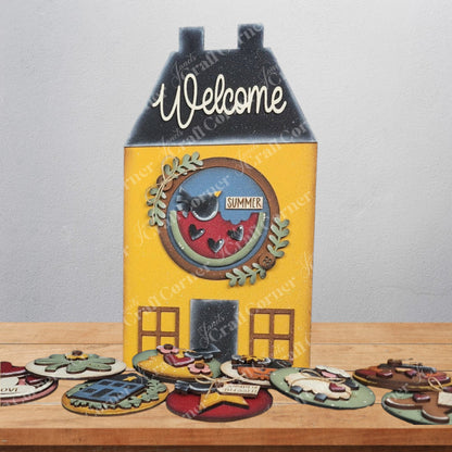 The Saltbox House Round Display by Janet's Craft Corner is a DIY seasonal home decor craft kit featuring a wooden saltbox house sign with "Welcome" on the roof and "Summer" in a circular window, plus colorful display inserts. Set of 1.