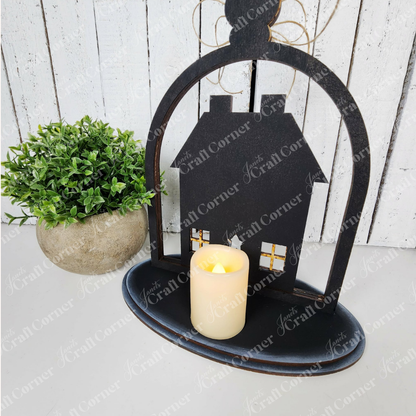 The Salt Box Votive Holder Cloche by Janet's Craft Corner—a DIY decor kit—features a votive holder on a black oval base with a flameless candle and a green potted plant, set against a white wooden background.