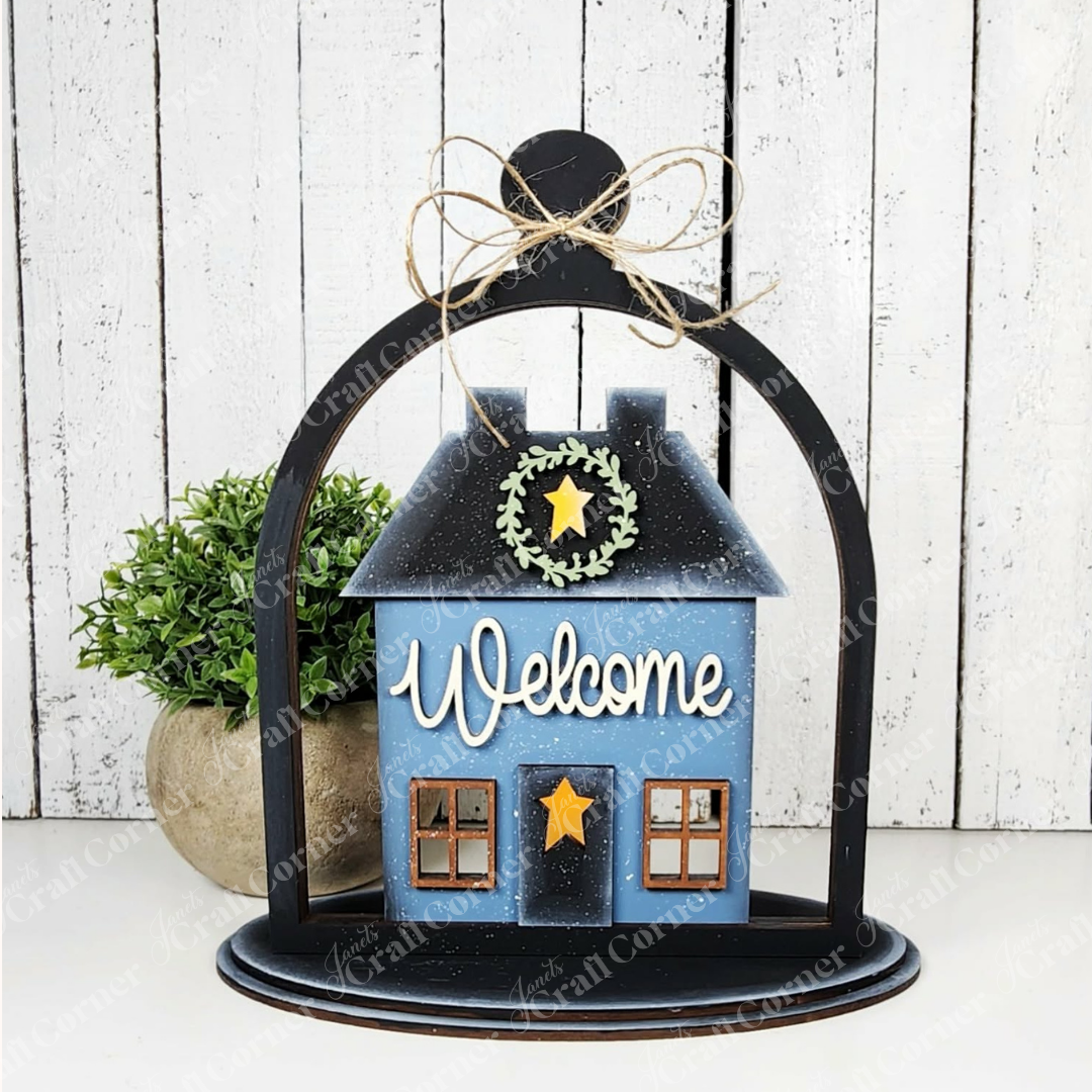 The Salt Box Votive Holder Cloche by Janet's Craft Corner is a blue DIY house kit with a “Welcome” sign, stars, twine bow top, green wreath accent, and cozy potted plant backdrop—perfect for charming seasonal home decor.