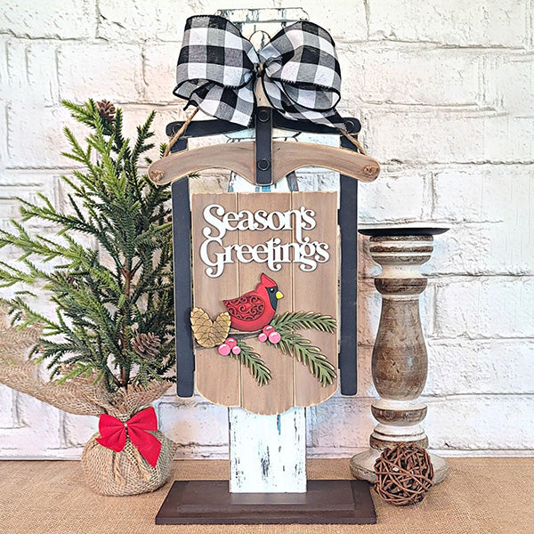 Create a festive holiday atmosphere with the Season's Greetings Sled Door Hanger from Janet's Craft Corner, featuring a red cardinal and pinecones. Adorned with a black-and-white checkered bow on top, this charming display includes a small burlap-wrapped pine tree on the left and a wooden candle holder on the right – perfect for your own DIY projects or as part of your seasonal decor.