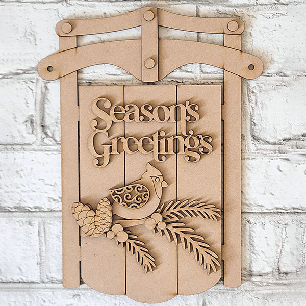 The Season's Greetings Sled Door Hanger from Janet's Craft Corner features carved "Season's Greetings," with a bird, pinecones, and branches on wood. It's an inviting DIY winter decor addition.