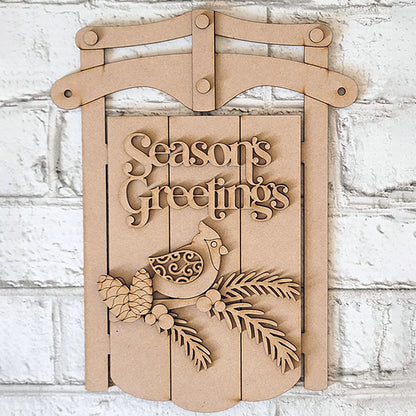 The Season's Greetings Sled Door Hanger from Janet's Craft Corner features carved "Season's Greetings," with a bird, pinecones, and branches on wood. It's an inviting DIY winter decor addition.